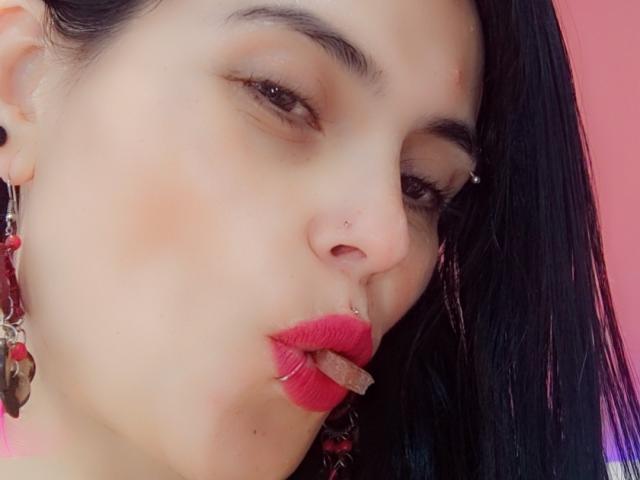 CharlotteCrofort's face with red lipstick, black earrings, nose piercing, and septum ring, looking down with slightly parted pink lips. She has dark hair and light skin.