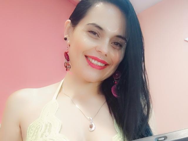 CharlotteCrofort with shiny skin, black hair, red lipstick, smiling, wearing pink earrings, silver necklace, lace top, and visible cleavage, in a pink room.