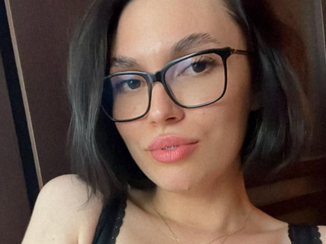MonaLiss with black glasses, pink lipstick, and black lace top, showing her face and slightly parted lips. She's got dark hair and a subtle smile. Boobs are partially visible.