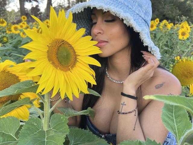 Stefeney Collin con un grande girasole giallo, che bacia i petali, indossa un cappello da pescatore in denim, una collana di perle, un braccialetto nero, mostra dei tatuaggi e ha un seno abbondante, in un campo di girasoli, con gli occhi chiusi, e sembra carina..