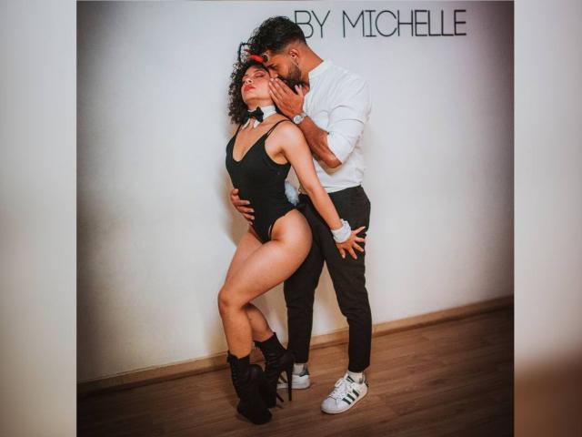IvannaAndFriend in a black one-piece, high heels, and wristbands, bent forward, hands on her neck. A man in a white shirt and black pants grips her.