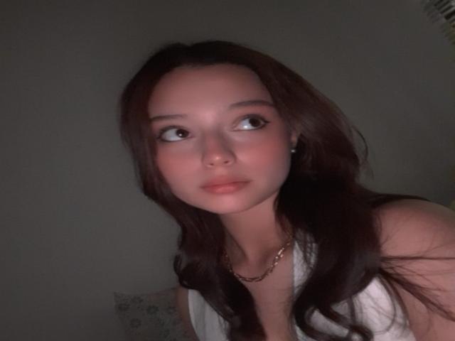 AngellMoore with long brown hair, wearing a white top, has a neutral expression and a gold necklace, looking to the side, with soft lighting on her face.