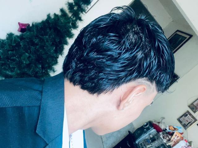 RichiMontes, com cabelo preto e brilhante, com aparência molhada, vestindo um blazer preto, camisa branca e um brinco, com a cabeça virada para o lado, mostrando a orelha e um penteado curto e ondulado..
