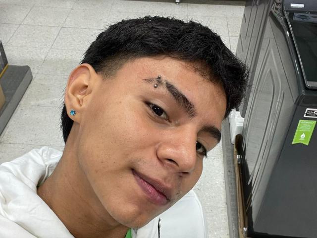 RichiMontes, con cabello negro corto y un pequeño piercing en la ceja derecha, vistiendo una sudadera blanca, sonriendo ligeramente. Tiene un arete azul y piel clara..