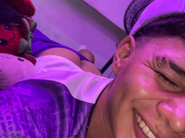 RichiMontes, con un piercing al naso, sorride, indossa una camicia viola e bianca e un cappello bianco, mostrando il sedere in biancheria intima viola, accanto a un giocattolo rosso e bianco di peluche..