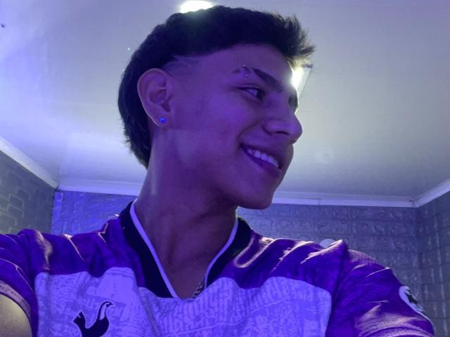 RichiMontes, sonriendo, lleva una camiseta de fútbol morada y blanca con un logo negro, tiene un piercing en la nariz y un corte de pelo a un lado con un pequeño moño, y pendientes azules..