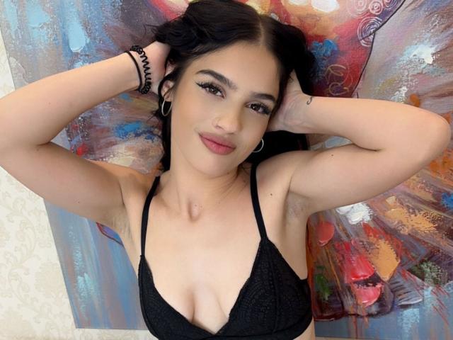 CherryDevine with black lace bra, small tits, armpit hair, arms raised, smiling, dark hair, hoop earrings, black bracelets, pink lipstick, and painted background.
