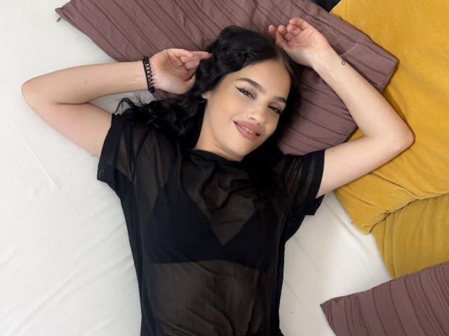 CherryDevine with black wavy hair, lying on a bed, arms above head, smiling, wearing a sheer black top, exposing her small tits, brown and yellow pillows.
