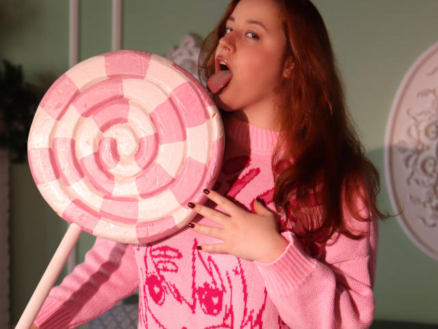PolinaGrace with red hair, tongue out, licking giant pink and white lollipop, wearing pink sweater with pink handprints, holding lollipop, pink nail polish, soft light.