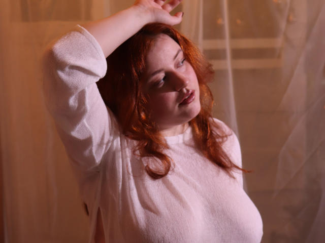 PolinaGrace with red hair, wearing a white, long-sleeve, textured shirt, arms raised, looking to the side, soft pink lighting, gentle expression, visible breasts, casual, relaxed pose.