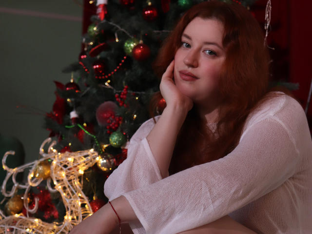 PolinaGrace with red hair, white sheer top, hand on cheek, sitting by a decorated Christmas tree with lights and ornaments, looking slightly to the side, soft smile.