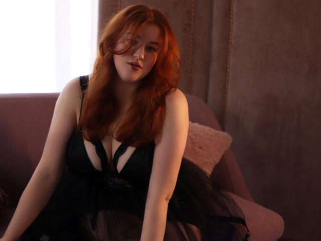 PolinaGrace with red hair, black lace dress, and cleavage, sitting on a purple sofa, looking at the camera with a subtle smile. Her tits are visible through the dress.