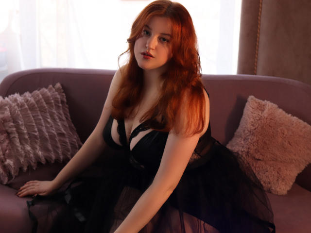 PolinaGrace with red hair, wearing a black lace dress, sitting on a purple couch, showing her tits and a cute face, with a pink fluffy pillow on the left.