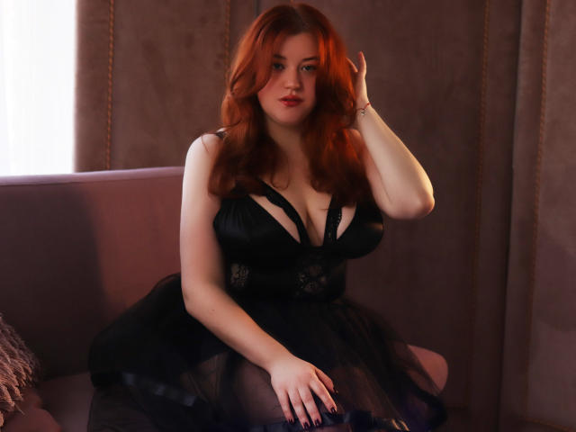 PolinaGrace with red hair, black lace dress, and sheer skirt, sitting on a pink sofa, hand in hair, showing large tits, and a sexy, confident expression.