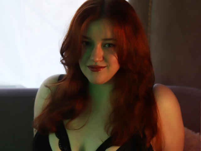 PolinaGrace with red hair, wearing black lace lingerie, has fair skin, small tits, and a subtle smile, sitting in soft green and blue light.