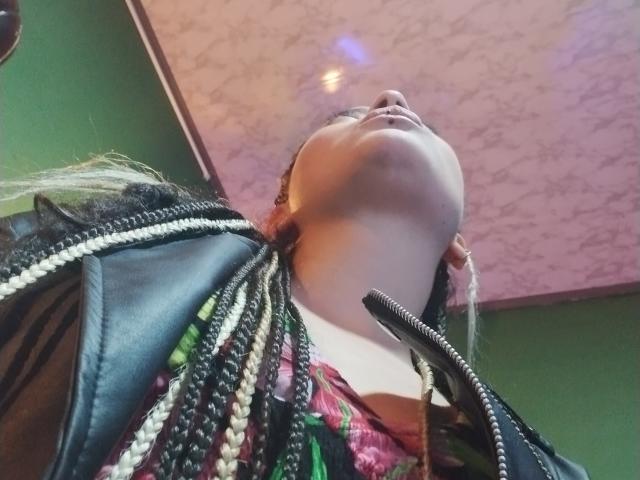 Low-angle shot of LunaTikka with braided hair, black leather jacket, and colorful top, head tilted back, showing her face and neck. Her pussy is not visible.
