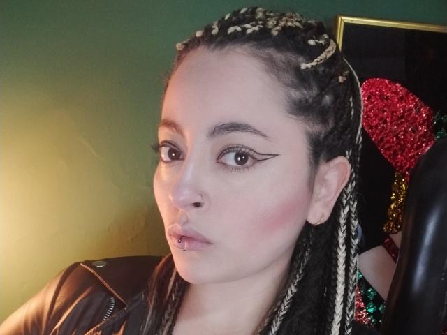 LunaTikka with braided hair, pale skin, and black eyeliner, has a nose piercing and a lip piercing, wearing a leather jacket, and a green wall in the background.