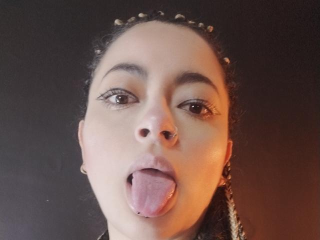 LunaTikka has braided hair, a nose ring, and a tongue piercing with her tongue out, looking directly at the camera with her brown eyes. Her lips are slightly parted.