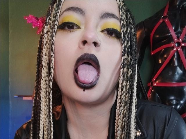 LunaTikka with braided blonde and black hair, yellow eyeshadow, black lipstick, tongue out, small nose piercing, wearing black leather, pink glitter hair accessory, and red harness in background.