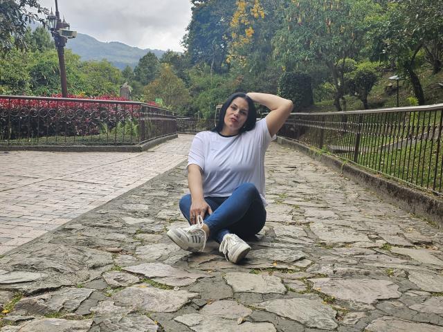 LizaRussell with black hair, white t-shirt, blue jeans, and white sneakers, sitting on a stone path, hand in hair, looking at camera, wooded background, cloudy sky.