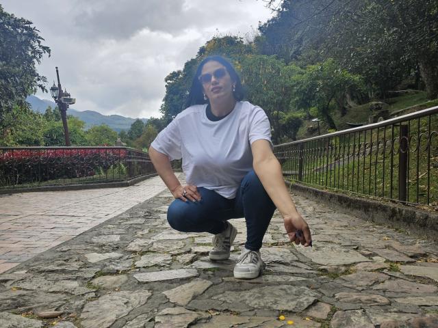 LizaRussell is squatting on a stone path, wearing a white t-shirt, blue jeans, and white sneakers, with her right hand on her knee and her left hand down.