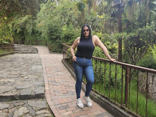 LizaRussell stands on a brick path, wearing a black sleeveless top, blue jeans, and white sneakers, with her arms on a metal railing, looking at the camera.