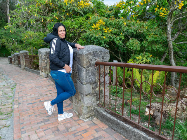 LizaRussell leans against a stone wall, wearing a black hoodie, blue jeans, and white sneakers, with her hand on her hip, and a slight smile on her face.