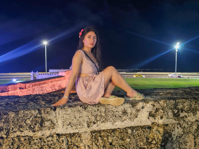JainaRogers with long brown hair, red flower, pink dress, and beige shoes, sitting on a rough stone ledge at night, legs crossed, under bright lights, looking slightly to the side.