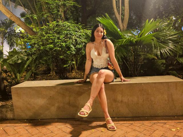 NicoColeman with tanned skin, dark hair, white lace top, denim shorts, and sandals, sitting on a concrete bench, legs crossed, green foliage in the background, sunlight filtering.