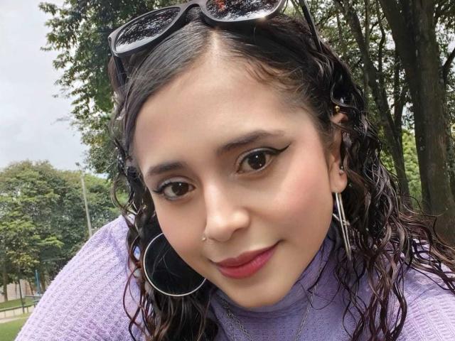 EliBaker has wet curly black hair, brown eyes, a nose ring, and is wearing a purple sweater with large hoop earrings. Sunglasses rest on her head. She's smiling.