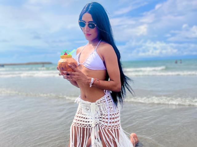 FallonRogers with long black hair, white bikini top, and white crocheted skirt, holding a coconut with a tiny pineapple on top, wearing sunglasses, standing on a beach.