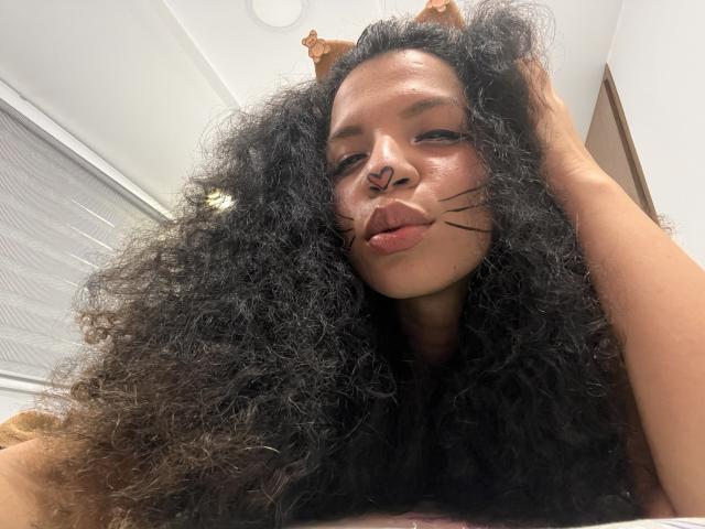 TiaraKitty with big curly black hair, brown skin, and a nose ring, wearing cat ears and black whisker makeup, pouting with a hand in her hair, looking down.