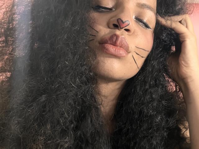 TiaraKitty has curly black hair, a heart on her nose, black cat whiskers, and pouty lips, with her hand touching her face, looking cute and slightly goofy.