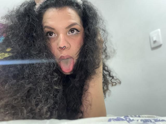 TiaraKitty with long curly black hair, nose septum piercing, and tongue out, wearing cat ears. She has big brown eyes and is looking at the camera.
