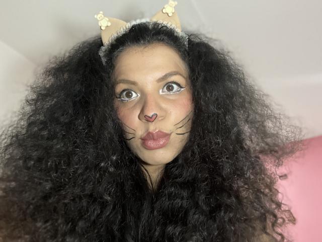 TiaraKitty with curly black hair, cat ears, and nose ring, pouting with painted whiskers and pink heart nose. Her tits are not visible, just her face.