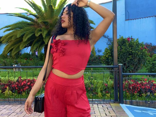 SophieSonyas with curly black hair, wearing a red crop top with flower accents and matching red pants, standing with one arm raised, holding a black purse.