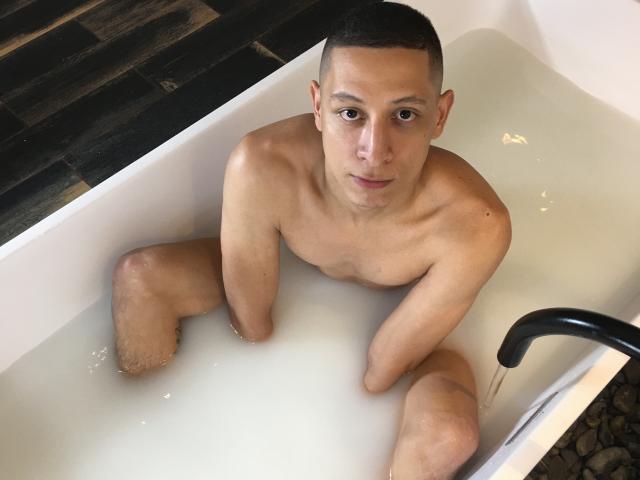 AdamCruz, naked, with short black hair, in a white bathtub, sitting in water, looking up with a neutral expression, showing a clean-shaven chest and a slightly visible, flaccid penis.