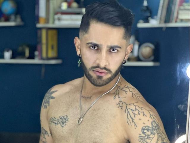 Shirtless ChristAnge with dark hair, beard, tattoos, and ear plugs, wearing a necklace, looking at camera, dark blue background, shelves with books and small items.