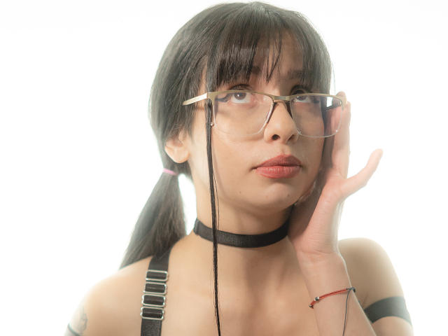 LilaBeauty with black pigtails, wearing glasses and a black choker, touching her face, has red lips, and a tattoo on her right shoulder, wearing a strap top.