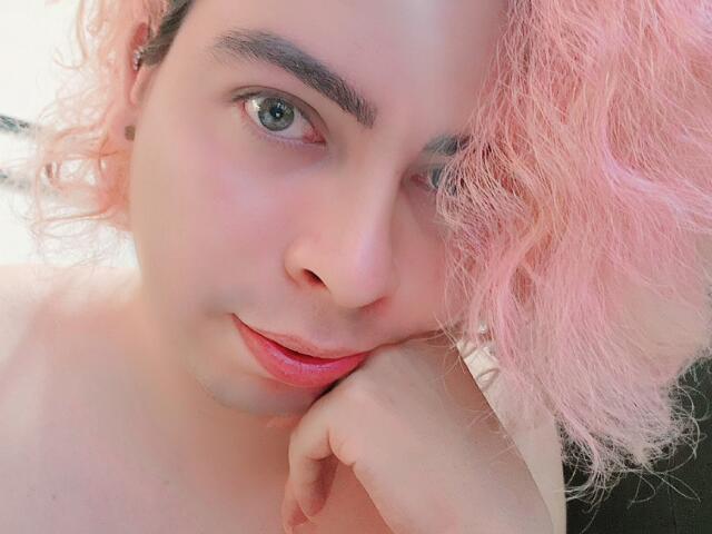 TsFoxCandy con cabello rizado rosa, piel clara, ojos verdes y labios rosados, con el mentón apoyado en la mano, mirando directamente a la cámara con una sonrisa sutil. Luz suave y natural..