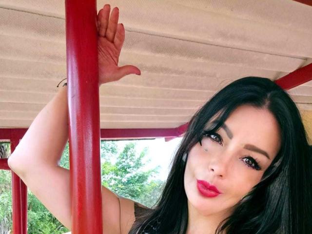 BrandyMostHottest with black hair, red lipstick, and eyeliner, holding a red pole, her hand above her head, looking at the camera with a slight smile.