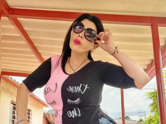 BrandyMostHottest with black hair, wearing black and pink top, black sunglasses, pink lipstick, adjusting sunglasses, standing under red and beige roof, showing her ass in tight jeans.