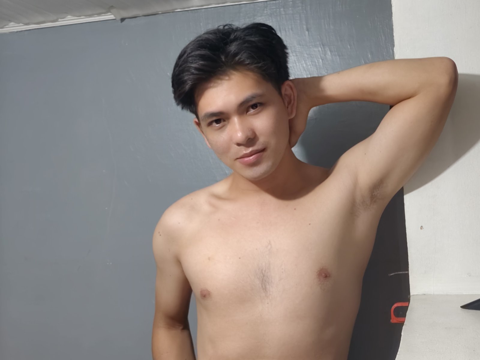 JimmyVortex, shirtless, with black hair, small nipples, and a slight smile, arm raised, showing armpit hair, standing against a gray wall.