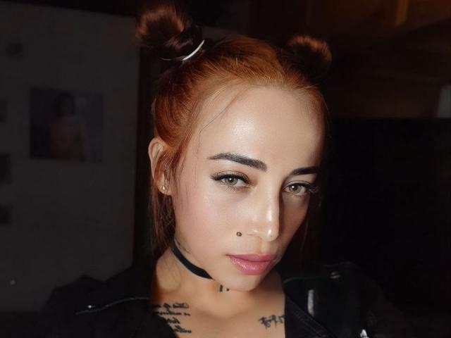 YeiSela ha la pelle color nocciola, i capelli rossi raccolti in due chignon, una collana nera, eyeliner, rossetto nero, un piercing al naso, un tatuaggio sul petto e un piccolo piercing al naso. Guarda verso la fotocamera..