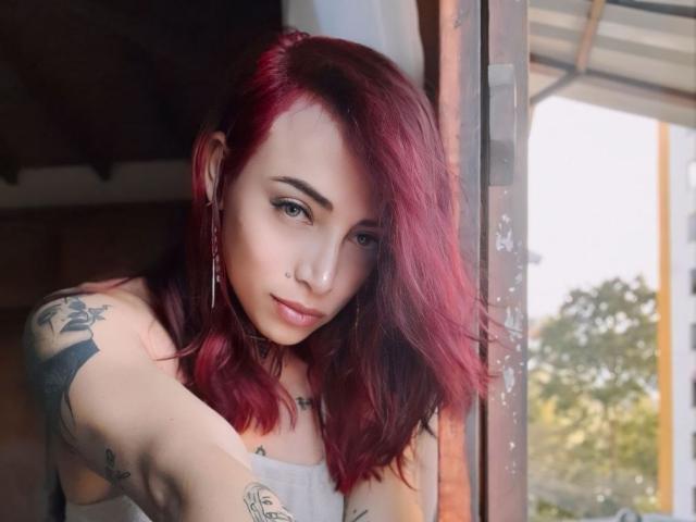 YeiSela with red hair, tattoos on arms, nose ring, and earrings, leaning on a window, looking down with a slight smile, sunlight in background, soft natural light.
