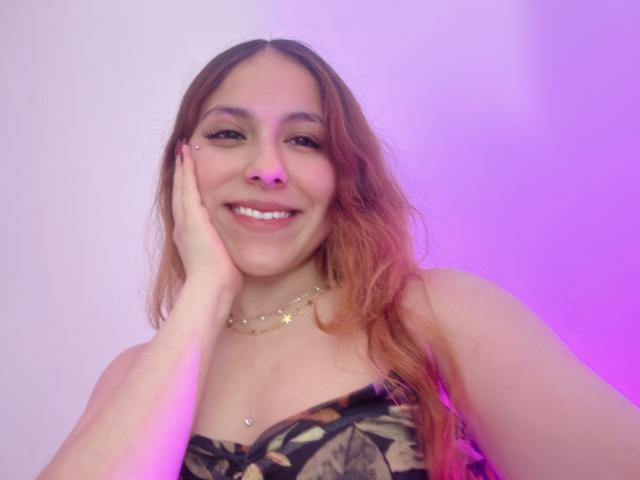 AmberlStha with long brown hair, smiling, hand on cheek, nose piercing, star necklace, and a patterned strapless top, showing her tits, in a pink-lit photo.