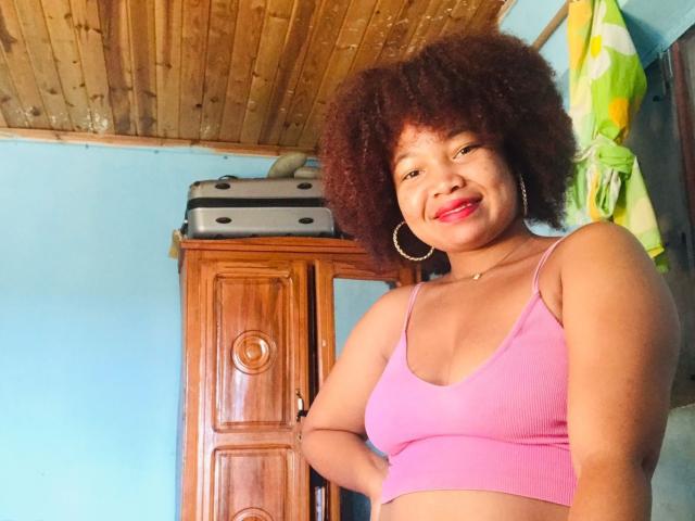 Orabelle has curly brown hair, red lipstick, and wears a pink sports bra, showing her small tits, with hoop earrings and a confident smile, standing in a wooden room.