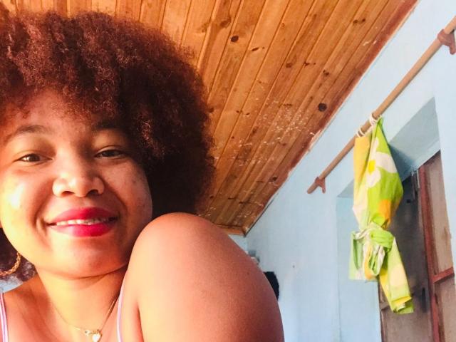 Orabelle has curly brown hair, red lipstick, and a warm smile. Her arm is raised, showing her shoulder. She's wearing a purple top with a wooden ceiling above.