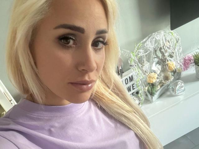 LunaSky, met blond haar, dikke wenkbrauwen en glanzende lippen, draagt een lila shirt, heeft een serieuze uitdrukking en toont een ingepakt cadeau in zilverfolie met gele rozen..