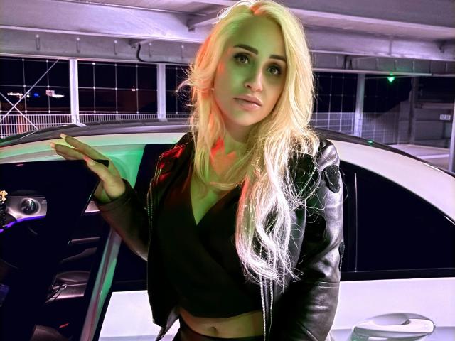 LunaSky-hot with long blonde hair, black leather jacket, and black top, standing by a futuristic car, hand raised, looking to the side with a slightly surprised expression.
